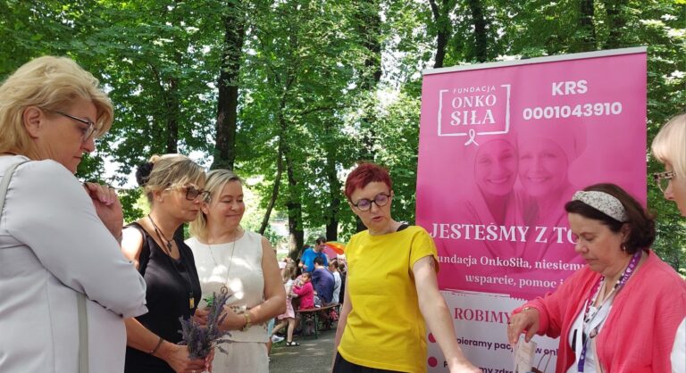 Plenerowa akcja profilaktyczna Fundacji ONKOSIŁA. Zdjęcie przedstawia grupę kobiet stojących przy stoisku Fundacji z edukacyjnymi modelami piersi.