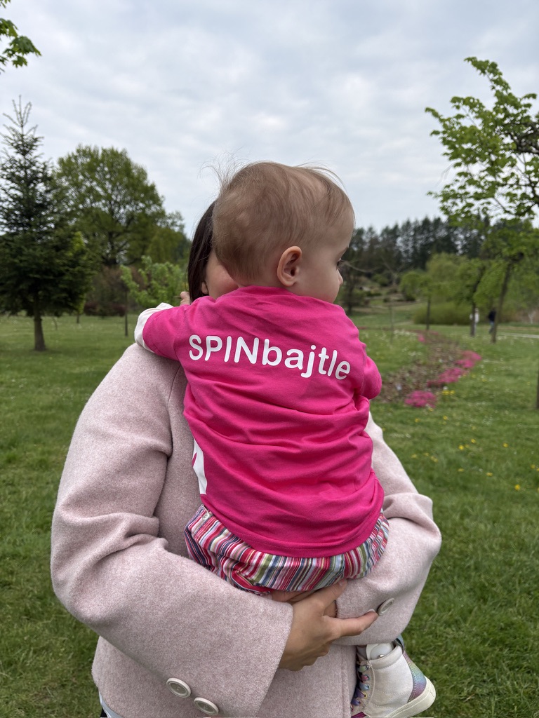 Dziecko w koszulce z napisem "SPINbajtle" trzymane na rękach przez kobietę.